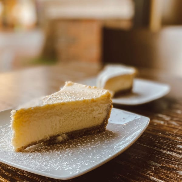Two slices of cheesecake with powdered sugar on white plates on a rustic wooden table.