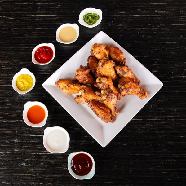 A white square plate of crispy chicken wings surrounded by six small bowls of dipping sauces.