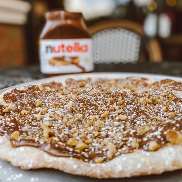 Nutella pizza with powdered sugar and chopped nuts, with a jar of Nutella in the background.
