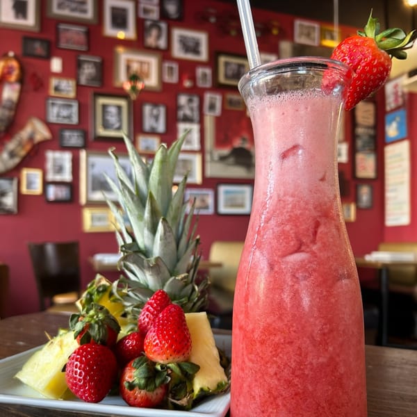 A strawberry smoothie in a tall glass with a straw, next to a plate of strawberries and pineapple.